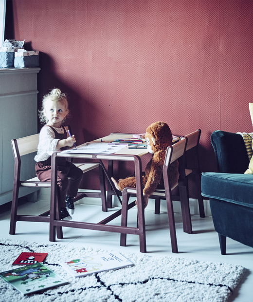 Малко момиченце седящо на детска маса в червена стая.