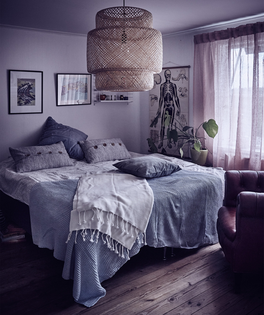 Уютна спалня със сиви стени и сиви и сини възглавници, хвърлени на легло, с дисяща над него лампа.