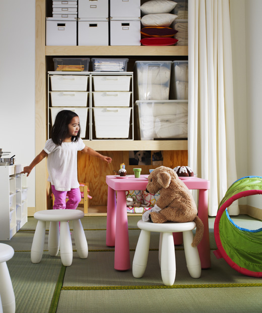 Дете организира чаено парти с плюшена играчка, на фона на отворени мебели за съхранение.