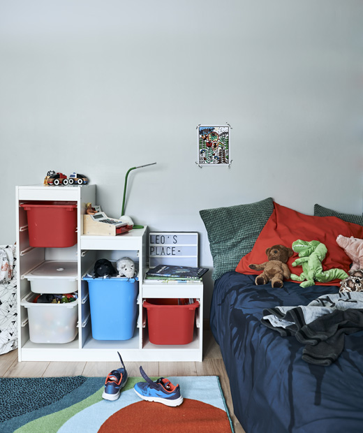Детска стая с цветни мебели за съхранение, килим и легло с възглавници и играчки.