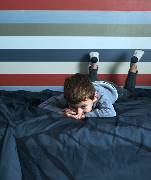 Момче лежи на легло с тъмносиня калъфка и шарен тапет.
